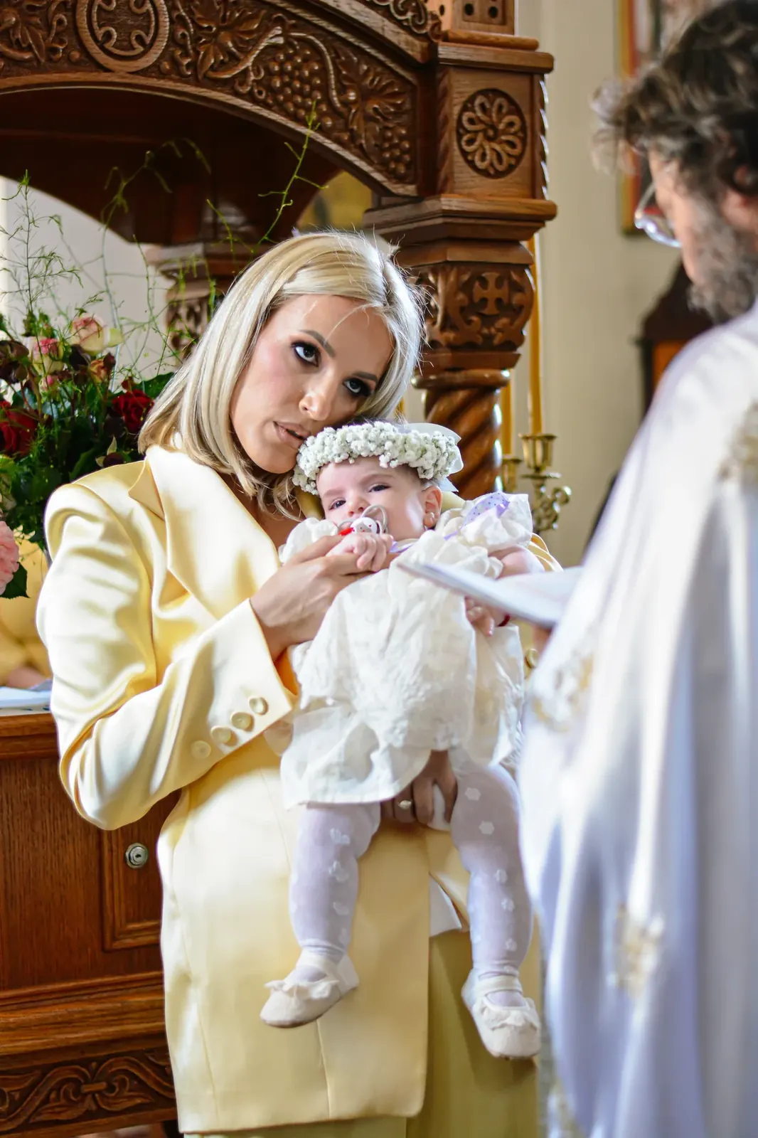 Mama drži bebu sa cvetnim vencem ispred ikonostasa